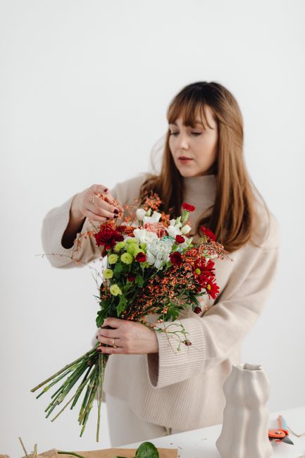 A young woman with long, light brown hair and bangs is arranging a vibrant bouquet of fresh flowers. The bouquet includes white, red, and green blooms, featuring carnations, chrysanthemums, and sprigs