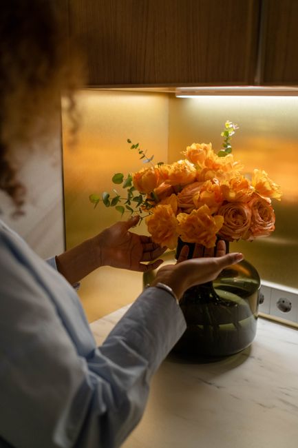A person in a light-colored shirt is arranging a bouquet of fresh flowers in a dark, round vase on a white marble surface. The bouquet features peach and pale pink roses, alongside orange carnations, 