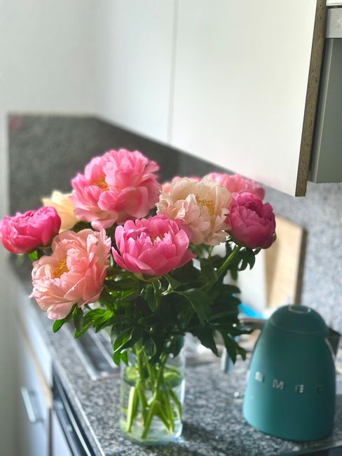 A bouquet of peonies in shades of pink, cream, and blush arranged in a clear glass vase placed on a kitchen countertop. The flowers have lush, ruffled petals with some blooms fully open and others sti