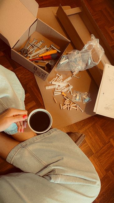 A person sitting on a wooden floor, holding a cup of black coffee in their left hand, with a floral arrangement kit on a large cardboard surface. The kit includes various flowers, foam blocks, scissor