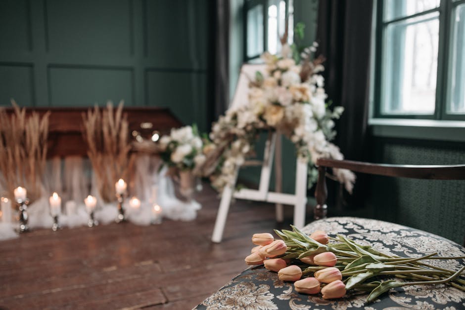 A floral arrangement scene featuring a collection of fresh, pale pink tulips with green stems and leaves resting on an ornate, patterned cushion on a wooden chair. In the background, a white floral sp