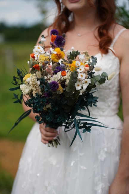 A woman wearing a white lace wedding dress with thin straps holding a large bouquet of fresh flowers. The bouquet includes purple, yellow, white, and orange blossoms such as daisies, chrysanthemums, a