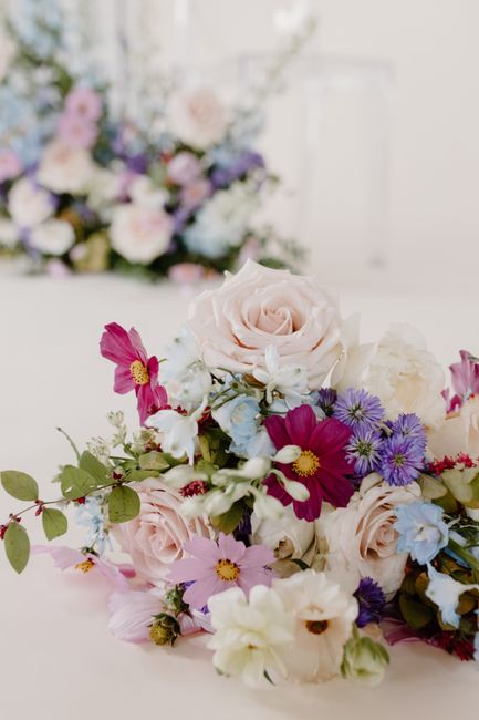 A close-up of a floral arrangement placed on a white surface, featuring a mix of pastel pink roses, white peonies, and sprigs of purple and violet asters, complemented by green foliage. The bouquet is